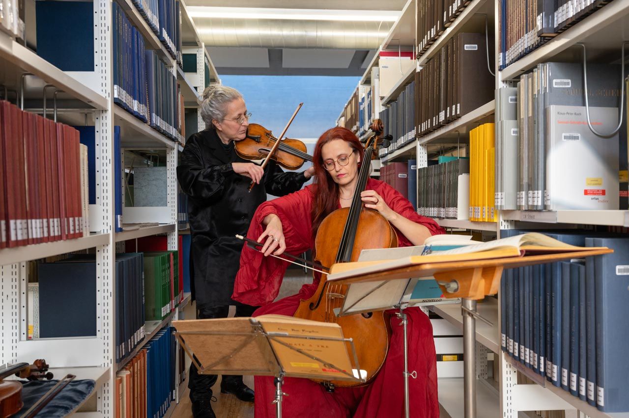 Zwei Frauen spielen Viola und Cello in einer Bibliothek, vor ihnen steht der Notenständer mit Musiknoten.