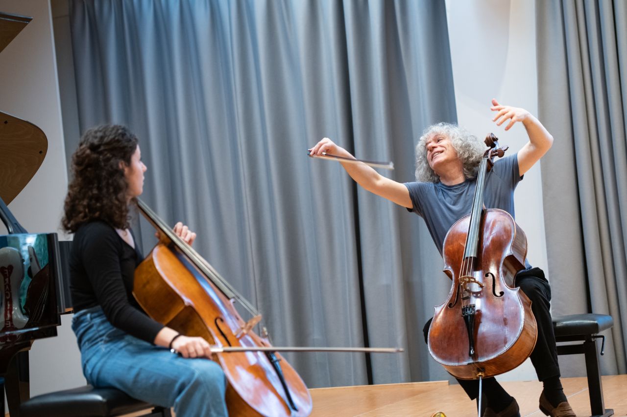 Der Cellist Steven Isserlis unterrichtet eine Studentin und macht, lachend, auslandende Armbewegungen.
