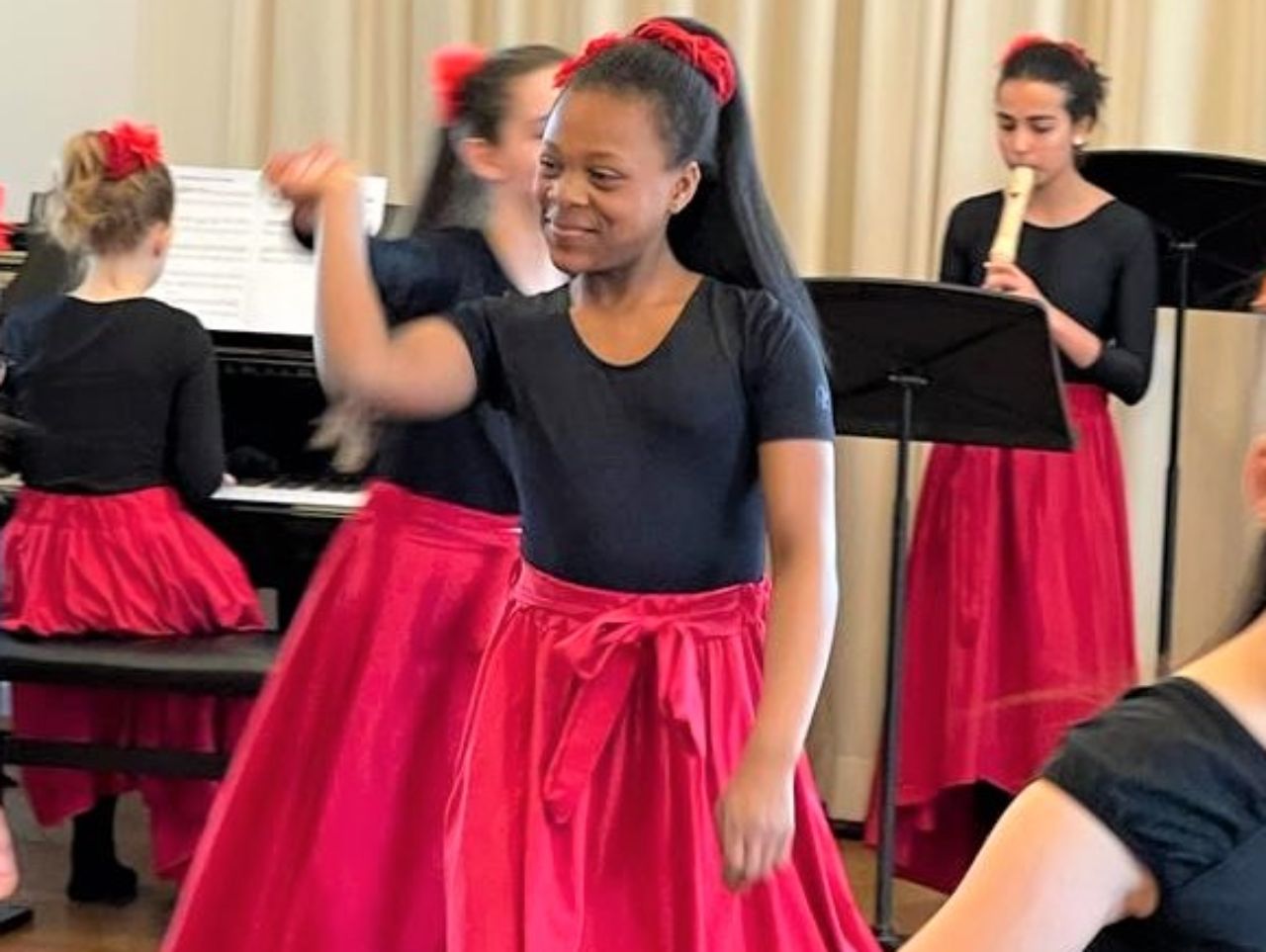 Ein Mädchen mit rotem Rock tanzt, im Hintergrund tanzen weitere Kinder. Jemand spielt Klavier, jemand Flöte.