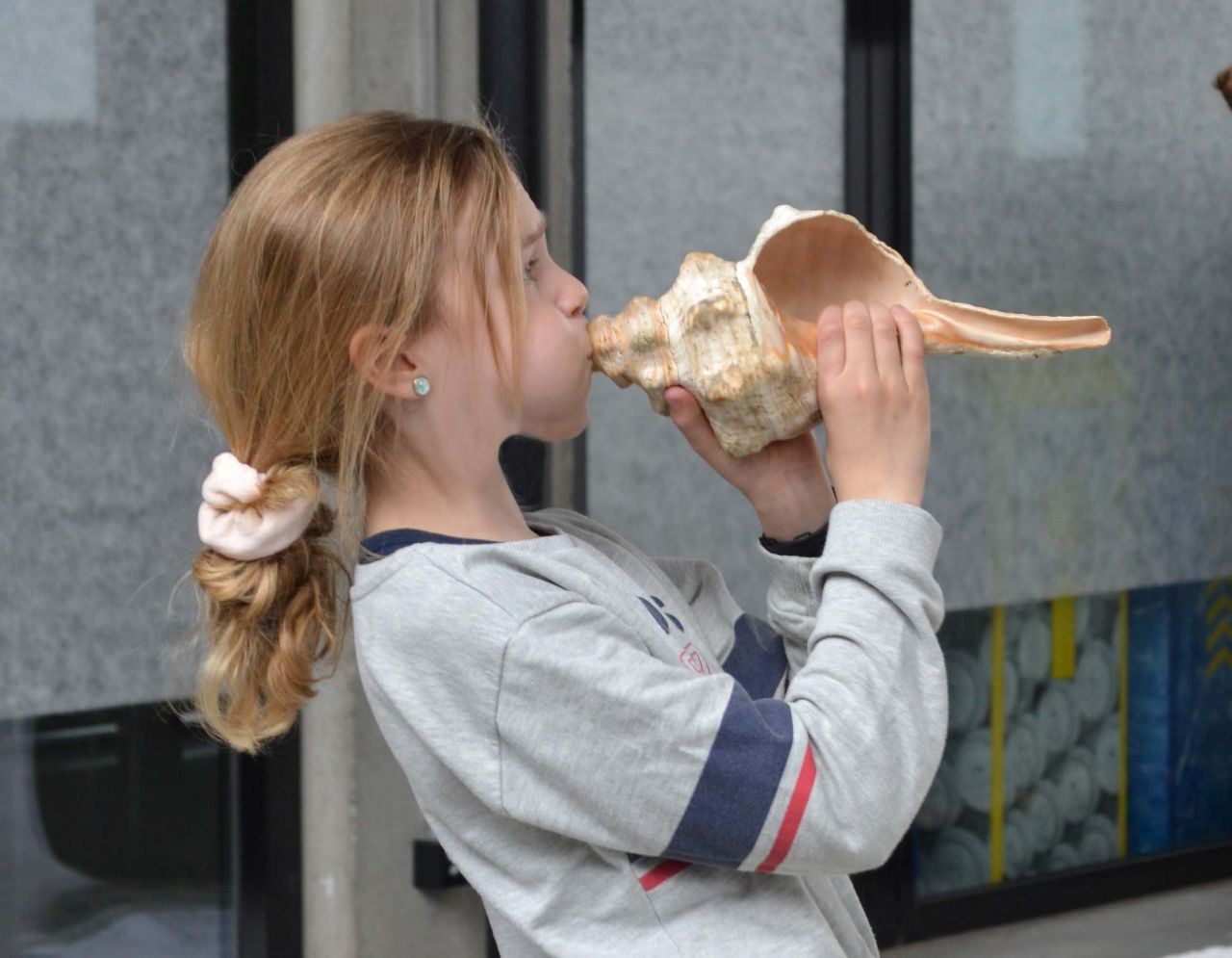 Ein Kind mit langen blonden Haaren, Jeans und einem grauen Pulli bläst in eine Muschel.