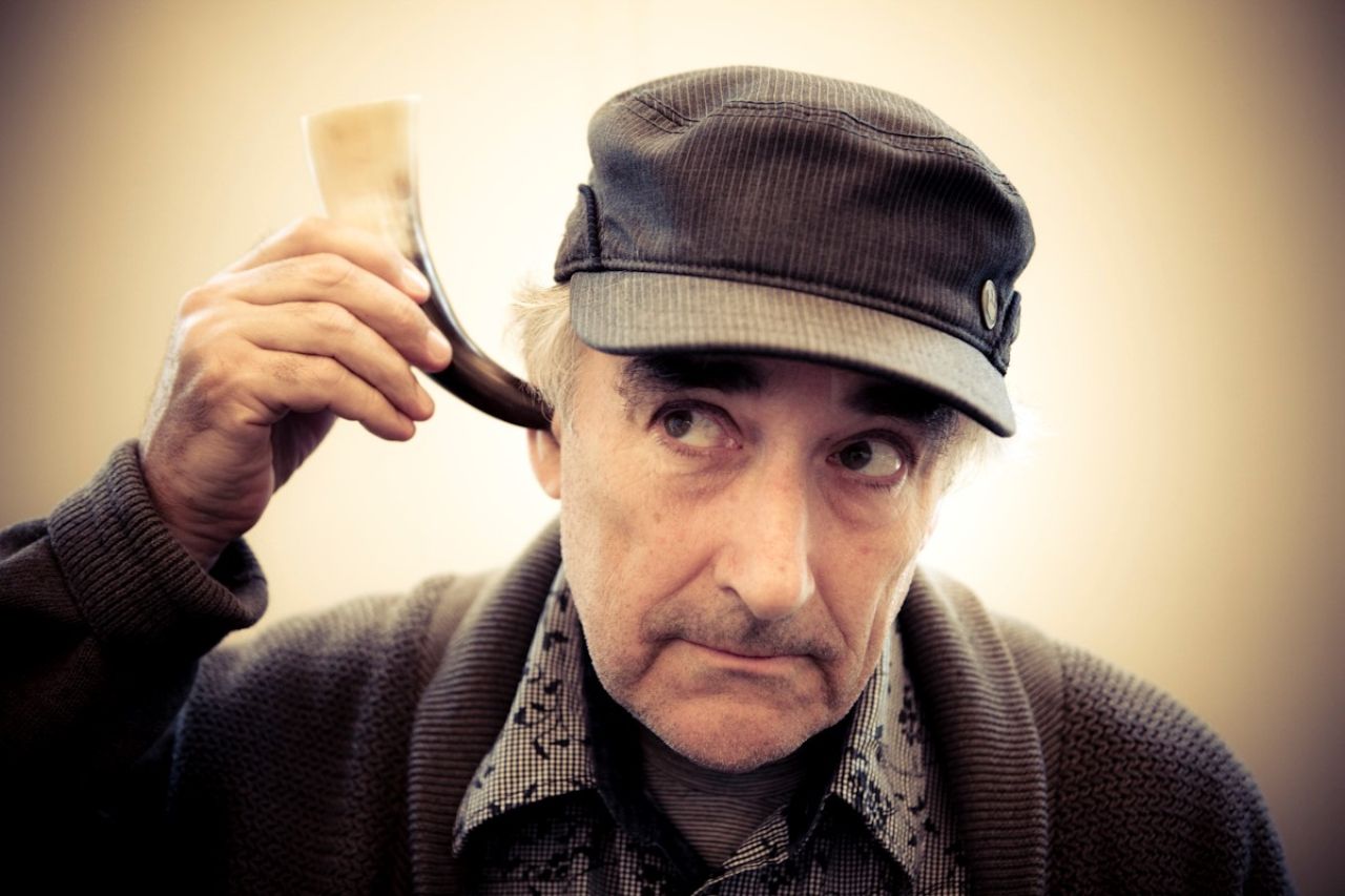 Ein Mann mit grauen Haaren hält ein Horn an sein Ohr, er trägt ein Beret und graue Kleidung.